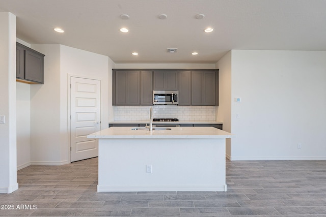 kitchen with gray cabinets, an island with sink, sink, light hardwood / wood-style flooring, and tasteful backsplash