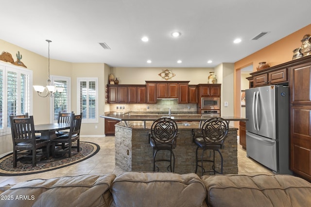 kitchen with an island with sink, dark stone countertops, decorative light fixtures, backsplash, and stainless steel appliances