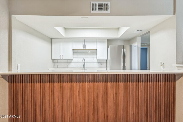 kitchen with kitchen peninsula, backsplash, stainless steel fridge, a raised ceiling, and white cabinetry