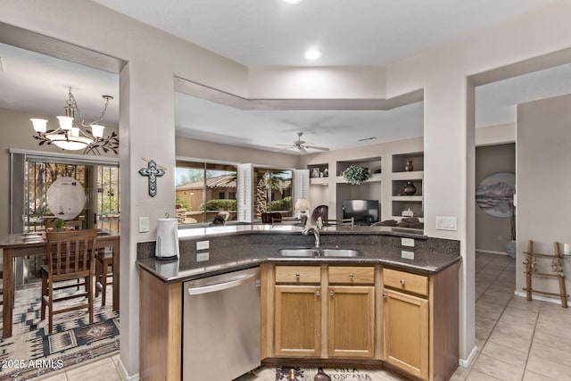 kitchen with built in shelves, dark stone counters, a sink, stainless steel dishwasher, and ceiling fan with notable chandelier