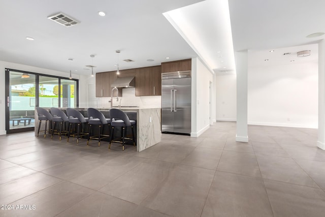 kitchen with an island with sink, built in refrigerator, a kitchen bar, decorative light fixtures, and range hood