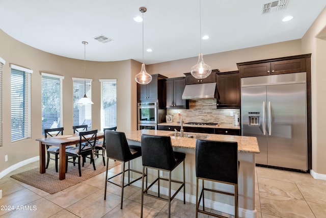 kitchen with light stone counters, a kitchen breakfast bar, an island with sink, pendant lighting, and stainless steel appliances