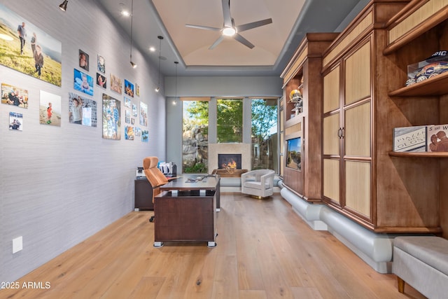 office area with a lit fireplace, a raised ceiling, light wood-style flooring, and a ceiling fan