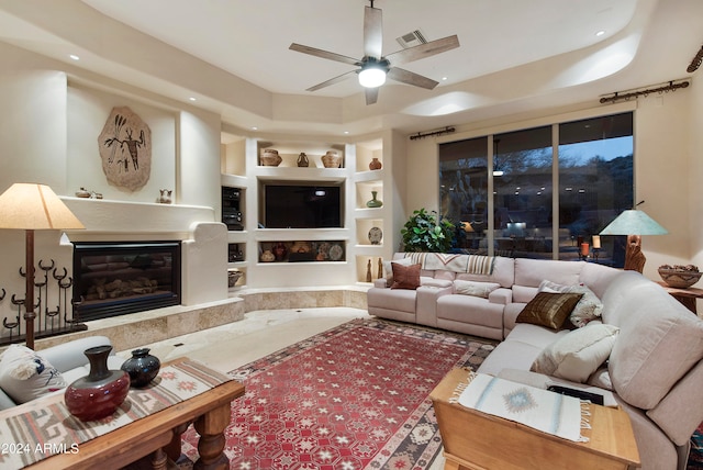 living room with a raised ceiling, built in shelves, and ceiling fan