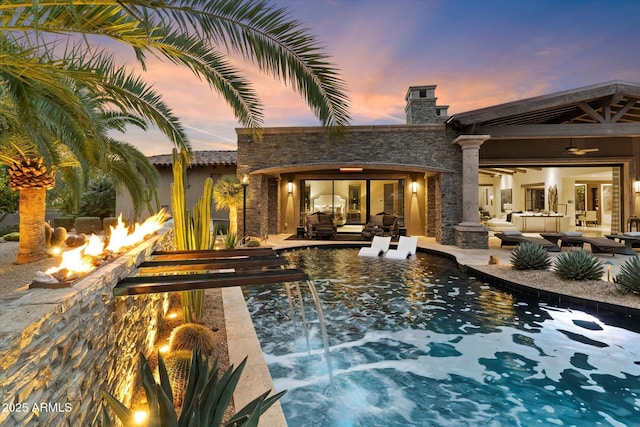 pool at dusk with a patio, pool water feature, an outdoor living space with a fire pit, and ceiling fan