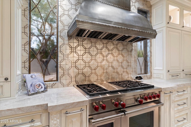 kitchen featuring double oven range, backsplash, cream cabinets, extractor fan, and light stone countertops