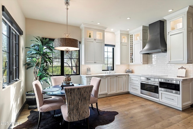 kitchen featuring wall chimney exhaust hood, white cabinetry, stainless steel oven, decorative light fixtures, and light hardwood / wood-style flooring