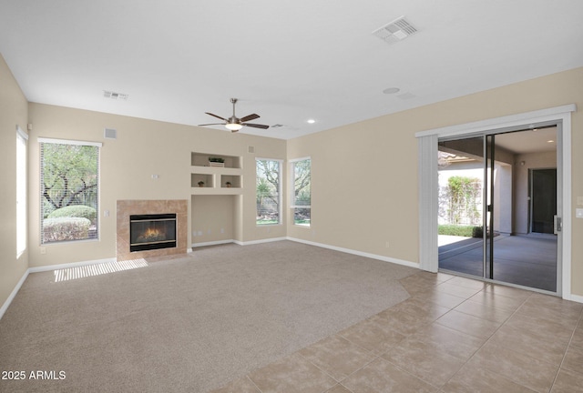 unfurnished living room with a tiled fireplace, ceiling fan, built in features, and light colored carpet