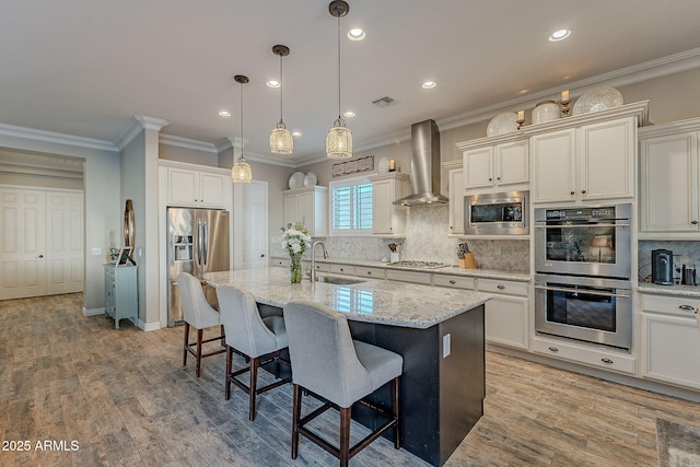 kitchen with wall chimney exhaust hood, sink, a center island with sink, appliances with stainless steel finishes, and pendant lighting