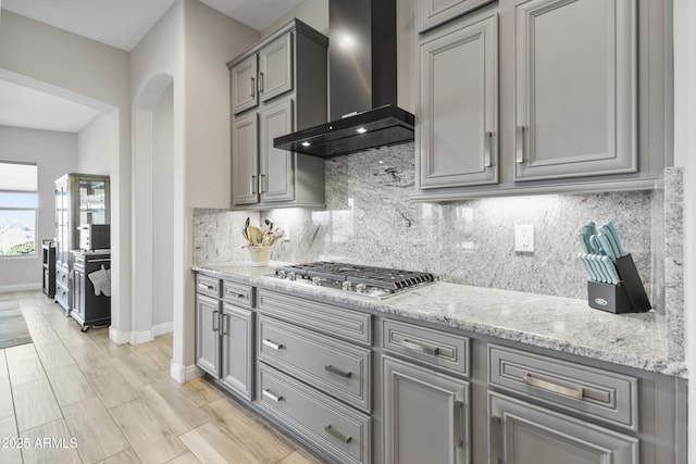 kitchen featuring arched walkways, gray cabinetry, wall chimney range hood, backsplash, and stainless steel gas stovetop