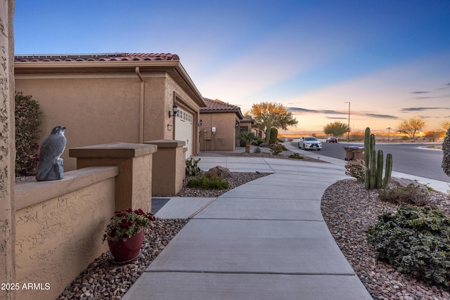 property exterior at dusk with a garage, a tile roof, driveway, and stucco siding