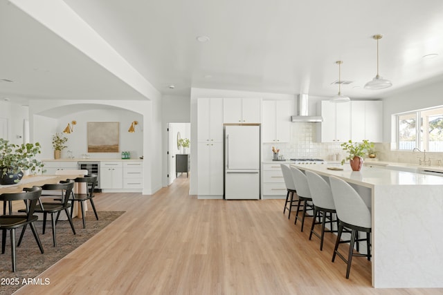 kitchen with wall chimney range hood, wine cooler, decorative light fixtures, white fridge, and white cabinetry