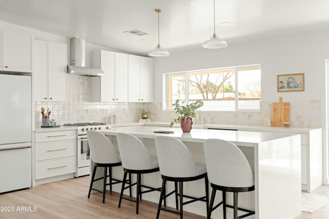 kitchen featuring wall chimney exhaust hood, white appliances, white cabinets, a kitchen island, and hanging light fixtures