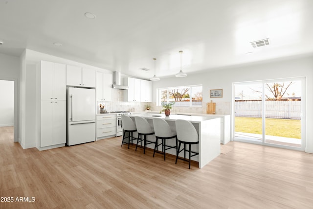 kitchen featuring white cabinets, white refrigerator, wall chimney range hood, high end stove, and decorative light fixtures