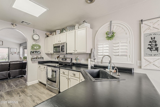kitchen featuring appliances with stainless steel finishes, a sink, arched walkways, dark countertops, and light wood-type flooring
