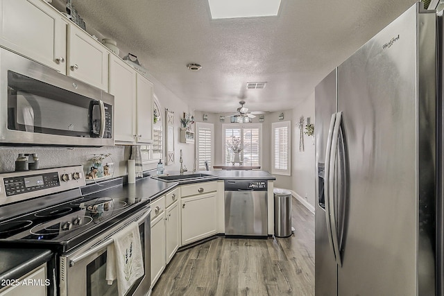 kitchen featuring dark countertops, a sink, a peninsula, light wood-style floors, and appliances with stainless steel finishes
