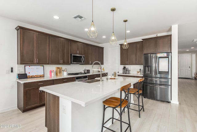 kitchen with backsplash, a kitchen island with sink, appliances with stainless steel finishes, decorative light fixtures, and dark brown cabinets