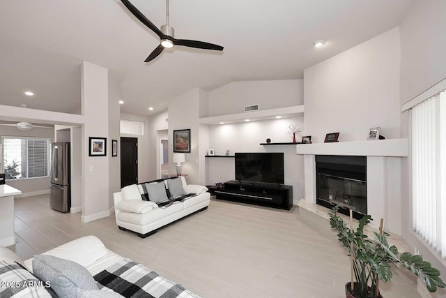 living area with visible vents, a ceiling fan, a glass covered fireplace, baseboards, and vaulted ceiling