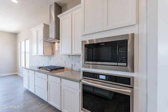 kitchen with stainless steel appliances, backsplash, white cabinetry, wall chimney exhaust hood, and light wood-type flooring