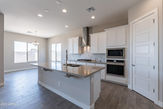 kitchen with an island with sink, wall chimney exhaust hood, white cabinetry, and appliances with stainless steel finishes