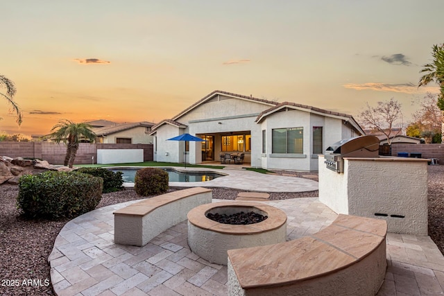 back house at dusk featuring an outdoor kitchen, an outdoor fire pit, a fenced in pool, and a patio