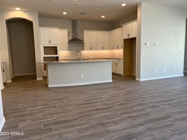 kitchen with an island with sink, wall chimney exhaust hood, hardwood / wood-style flooring, and white cabinetry