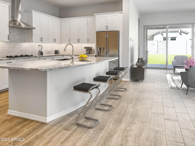 kitchen with an island with sink, stainless steel fridge, sink, wall chimney range hood, and light hardwood / wood-style floors