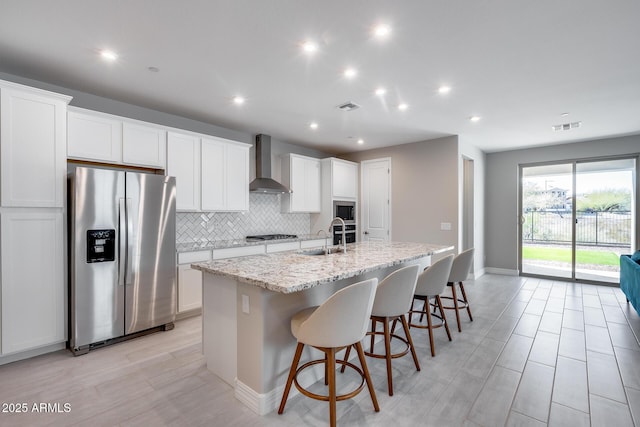 kitchen with wall chimney range hood, a kitchen island with sink, visible vents, appliances with stainless steel finishes, and white cabinetry