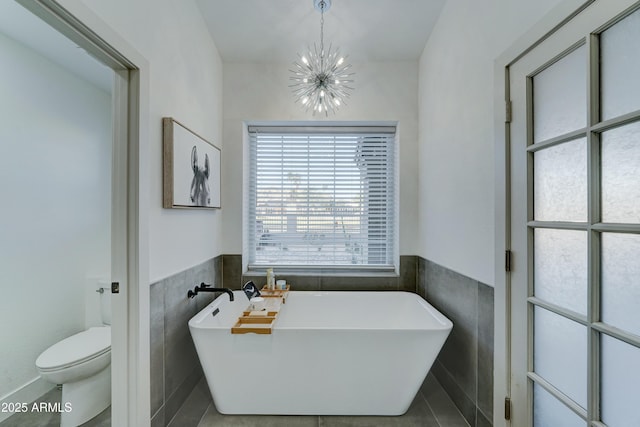 full bathroom with toilet, a wainscoted wall, an inviting chandelier, a freestanding bath, and tile walls