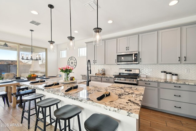 kitchen featuring appliances with stainless steel finishes, gray cabinets, hanging light fixtures, and a center island with sink