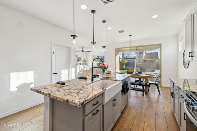 kitchen featuring gray cabinets, light stone countertops, stainless steel range oven, a center island with sink, and decorative light fixtures