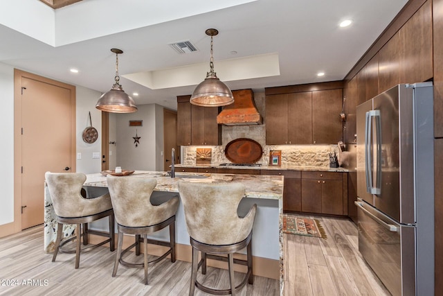 kitchen with hanging light fixtures, custom range hood, light hardwood / wood-style floors, dark brown cabinetry, and stainless steel appliances