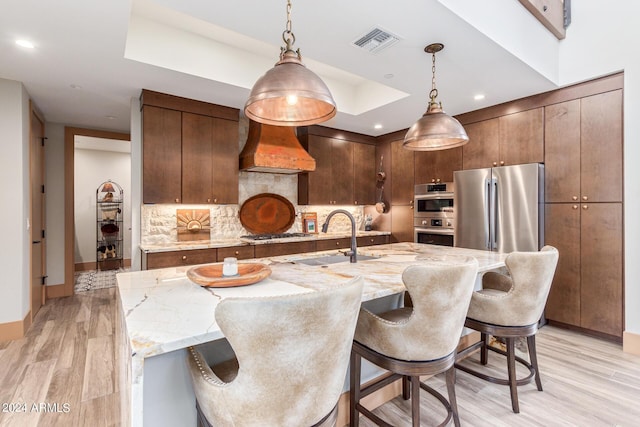 kitchen featuring a center island with sink, stainless steel appliances, custom range hood, and light hardwood / wood-style floors