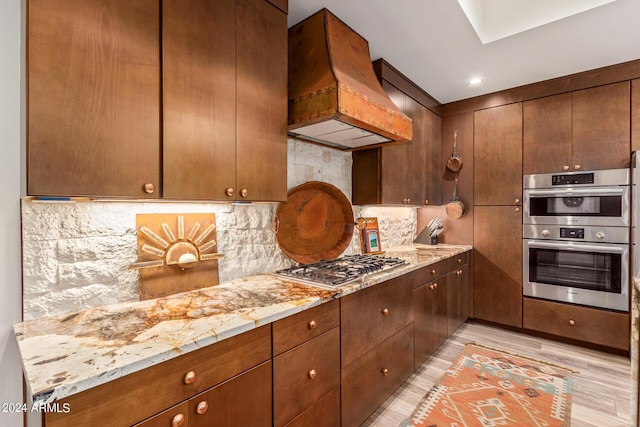 kitchen featuring appliances with stainless steel finishes, tasteful backsplash, light stone counters, custom exhaust hood, and light hardwood / wood-style floors