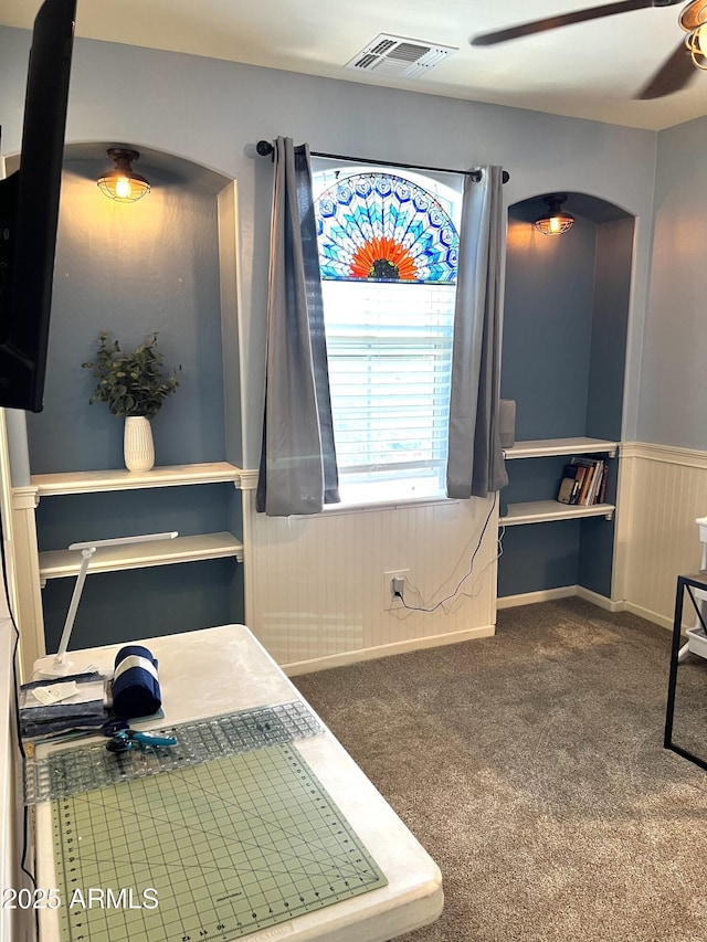 office space featuring a ceiling fan, dark colored carpet, a wainscoted wall, and visible vents