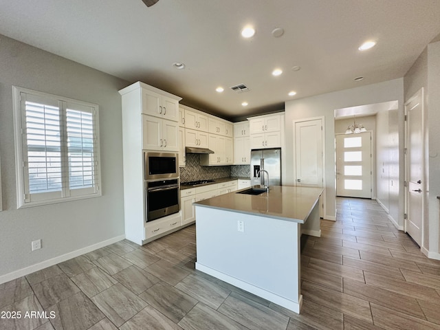 kitchen with tasteful backsplash, a chandelier, appliances with stainless steel finishes, a kitchen island with sink, and white cabinets