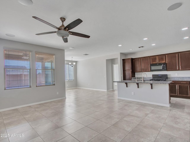 kitchen featuring a breakfast bar area, light stone counters, range, a center island with sink, and ceiling fan with notable chandelier