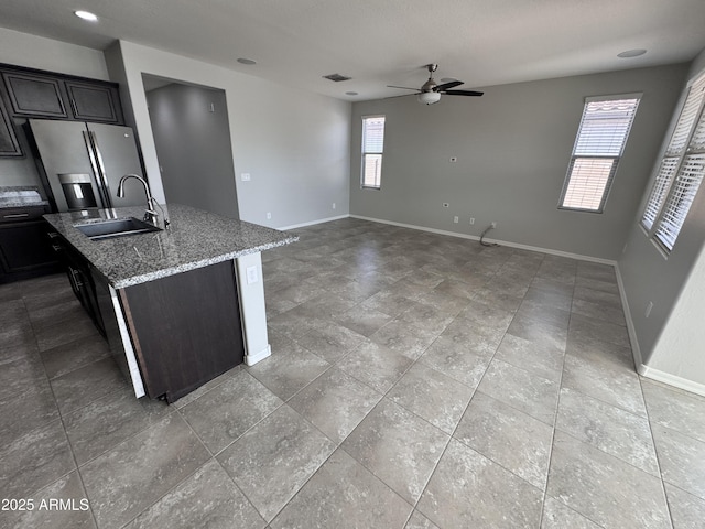 kitchen with sink, stainless steel fridge, ceiling fan, light stone counters, and a center island with sink