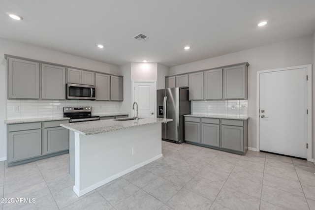 kitchen with a center island with sink, sink, gray cabinets, light stone counters, and stainless steel appliances