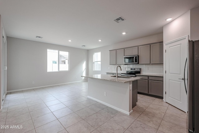 kitchen with backsplash, a kitchen island with sink, sink, appliances with stainless steel finishes, and light stone counters