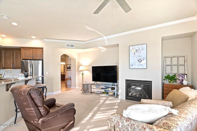 carpeted living room featuring ceiling fan, crown molding, a tiled fireplace, and sink