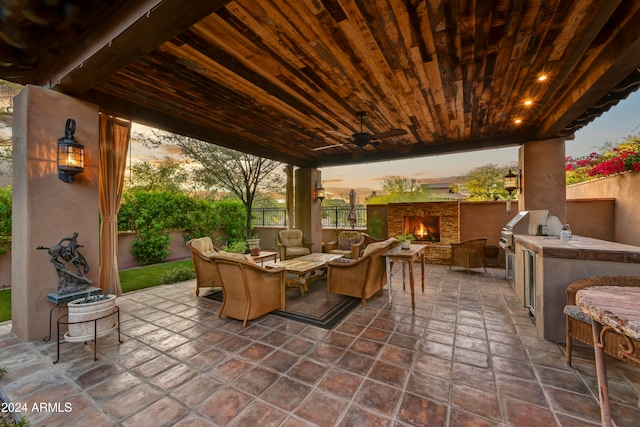 patio terrace at dusk with ceiling fan, area for grilling, and an outdoor stone fireplace