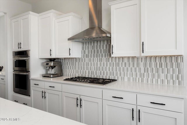 kitchen with white cabinets, appliances with stainless steel finishes, wall chimney exhaust hood, decorative backsplash, and light stone counters