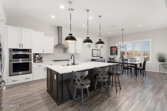 kitchen with wall chimney exhaust hood, white cabinets, a center island with sink, and hanging light fixtures