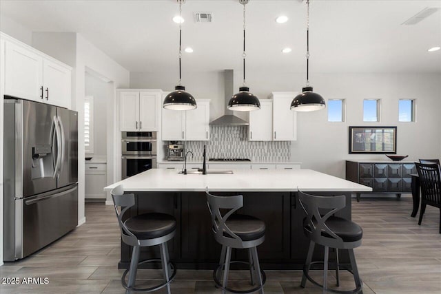 kitchen with hanging light fixtures, stainless steel appliances, wall chimney exhaust hood, and an island with sink