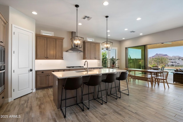 5724 E Scarlet St, Mesa, AZ 85215 - kitchen with wall chimney range hood, a breakfast bar area, a center island with sink, light countertops, and light wood-style flooring