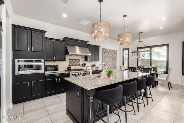 5212 S Seton Ct, Gilbert, AZ 85298 - kitchen with under cabinet range hood, a sink, dark cabinetry, an island with sink, and tasteful backsplash