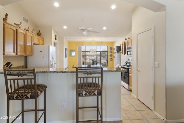 kitchen with light tile patterned flooring, a breakfast bar area, dark stone countertops, kitchen peninsula, and white appliances