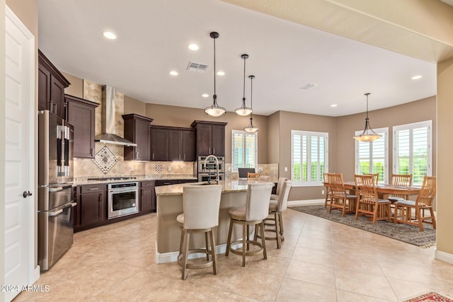 kitchen featuring appliances with stainless steel finishes, light stone counters, wall chimney exhaust hood, a kitchen island with sink, and decorative light fixtures