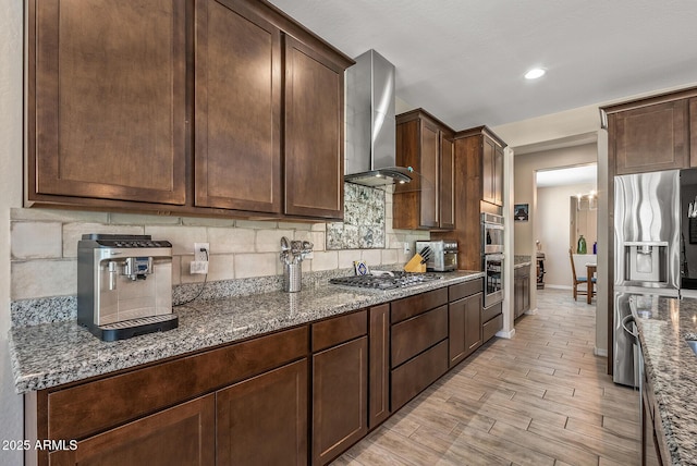 kitchen featuring stainless steel appliances, decorative backsplash, wall chimney range hood, and light stone counters